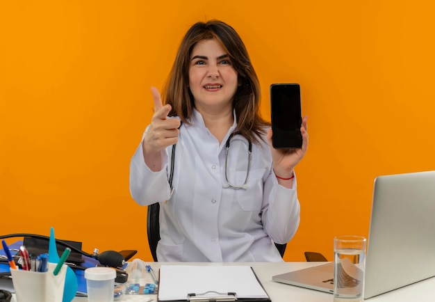 Unzufriedene Ärztin mittleren Alters, die medizinische Robe mit Stethoskop trägt, das am Schreibtisch sitzt, arbeitet auf Laptop mit medizinischen Werkzeugen, die Telefon halten und Ihnen Geste auf orange Wand zeigen