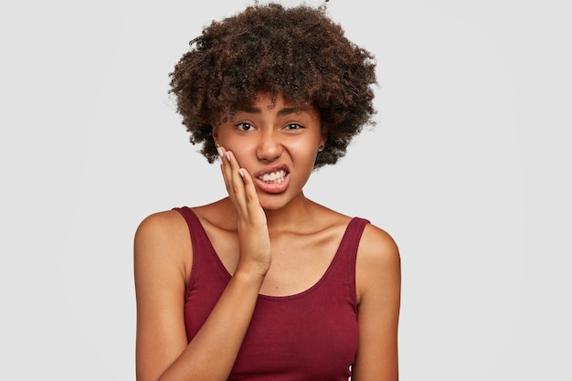 Unzufriedene schöne Afroamerikanerin hält Hand auf Wangen, fühlt Zahnschmerzen, biss die Zähne vor Schmerzen zusammen, hat dunkle, gesunde Haut und knackiges Haar, lässig gekleidet. Negative Gesichtsausdrücke