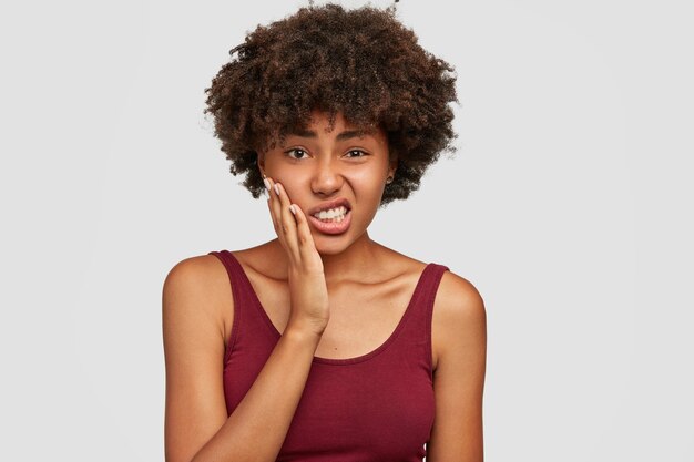 Unzufriedene schöne Afroamerikanerin hält Hand auf Wangen, fühlt Zahnschmerzen, biss die Zähne vor Schmerzen zusammen, hat dunkle, gesunde Haut und knackiges Haar, lässig gekleidet. Negative Gesichtsausdrücke