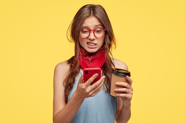 Unzufriedene schöne Frau schaut mit Ekel und Abneigung auf Handy, bearbeitet Foto in spezieller App, trinkt Kaffee zum Mitnehmen, posiert über gelber Wand, fühlt sich nicht mögen, verbunden mit Highspeed-Internet
