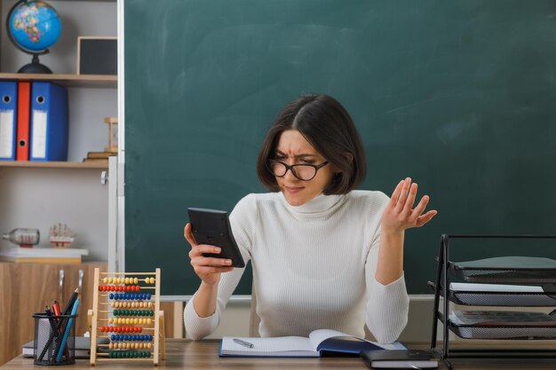 unzufriedene sich ausbreitende Hände junge Lehrerin mit Brille, die einen Taschenrechner hält, der am Schreibtisch mit Schulwerkzeugen im Klassenzimmer sitzt