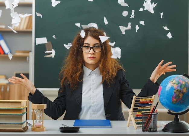 unzufriedene sich ausbreitende Hände junge Lehrerin mit Brille reißt Papier, das am Schreibtisch mit Schulwerkzeugen im Klassenzimmer sitzt