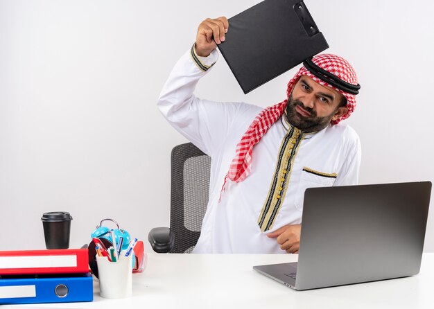 Unzufriedener arabischer Geschäftsmann in der traditionellen Kleidung, die am Tisch mit Laptop-Computer hält Zwischenablage mit negativen Emotionen sitzt im Büro sitzt