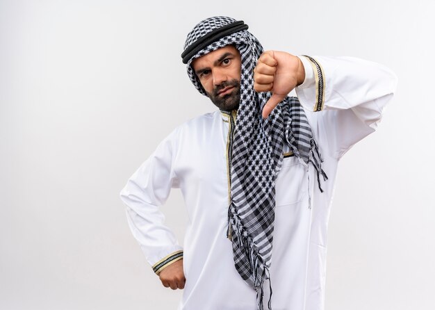 Unzufriedener arabischer Mann in traditioneller Kleidung, die Daumen nach unten stehend über weißer Wand zeigt