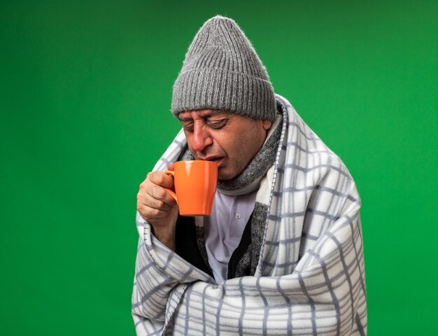 unzufriedener erwachsener kranker kaukasischer Mann mit Schal um den Hals, der eine Wintermütze trägt, die in kariertes Halten gewickelt ist und die Tasse isoliert auf grüner Wand mit Kopienraum betrachtet