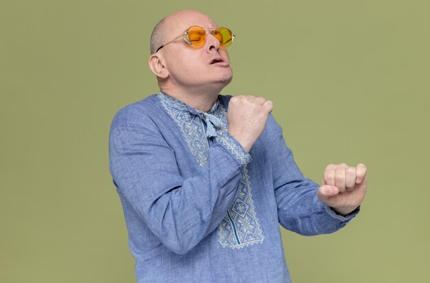 Unzufriedener erwachsener slawischer Mann in blauem Hemd mit Sonnenbrille, der mit geschlossenen Augen steht und vorgibt, etwas zu halten