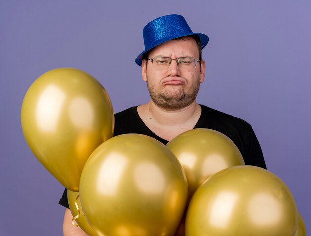 Unzufriedener erwachsener slawischer Mann in optischer Brille mit blauem Partyhut hält Heliumballons