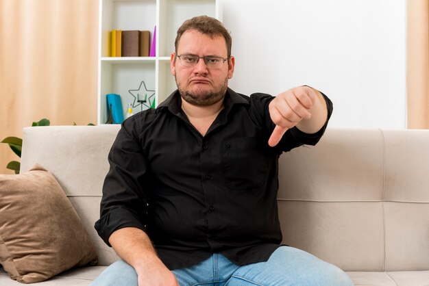 Unzufriedener erwachsener slawischer Mann in optischer Brille sitzt auf Sessel Daumen unten im Wohnzimmer