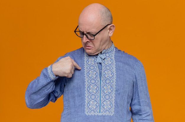 Unzufriedener erwachsener slawischer Mann mit optischer Brille, der auf sein blaues Hemd schaut und zeigt