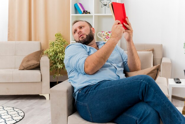 Unzufriedener erwachsener slawischer Mann sitzt auf Sessel, hält Buch und schaut zur Seite im Wohnzimmer