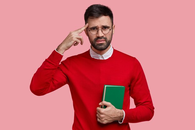 Unzufriedener europäischer Mann hält Finger an Schläfe, trägt roten Pullover, trägt grünes Lehrbuch, hat unglücklichen Gesichtsausdruck