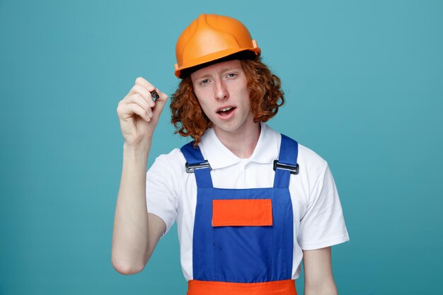 Unzufriedener junger Baumeister in Uniform, der einen Markierungsstift in die Kamera hält, isoliert auf blauem Hintergrund