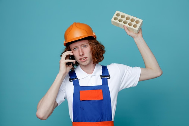 Unzufriedener junger Baumeister in Uniform, der Ziegel hält und am Telefon spricht, isoliert auf blauem Hintergrund