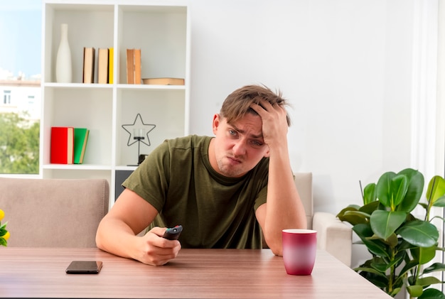 Unzufriedener junger blonder gutaussehender Mann sitzt am Tisch mit Telefon und Tasse, die Hand auf die Stirn hält, die Fernbedienung des Fernsehers hält