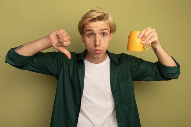 Unzufriedener junger blonder Kerl, der grünes T-Shirt trägt, das Tasse oben Seite unten hält Daumen unten zeigt