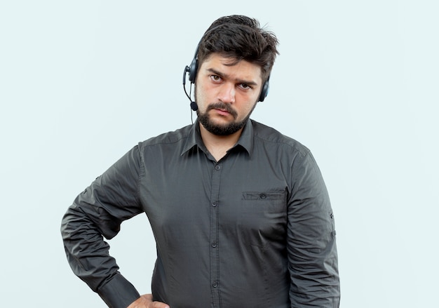 Unzufriedener junger Callcenter-Mann, der Headset trägt, das Hand auf Hüfte lokalisiert auf Weiß setzt