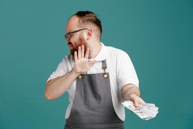 Unzufriedener junger Friseur in Uniform und Brille, der sich von Kopf zu Seite dreht und Geld in Richtung Kamera streckt, ohne Geste mit geschlossenen Augen, isoliert auf blauem Hintergrund