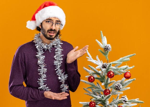 unzufriedener junger gutaussehender Kerl, der in der Nähe des Weihnachtsbaums steht und einen Weihnachtshut mit Girlande an den Halspunkten am Baum trägt, der auf der orangefarbenen Wand isoliert ist?