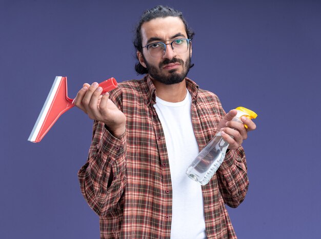 Unzufriedener junger gutaussehender Putzmann mit T-Shirt mit Moppkopf mit Sprühflasche isoliert auf blauer Wand