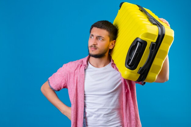 Unzufriedener junger gutaussehender Reisender, der Koffer hält, der mit stirnrunzelndem Gesicht über blauem Hintergrund beiseite schaut