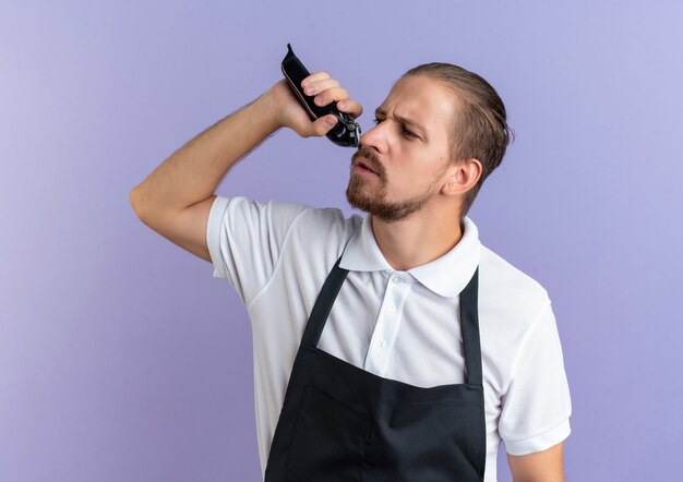 Unzufriedener junger hübscher Friseur, der Uniform hält, die Haarschneidemaschinen hält, die seinen eigenen Bart beschneiden, der Seite betrachtet, die auf lila Hintergrund lokalisiert wird