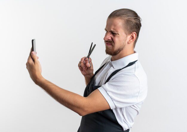 Unzufriedener junger hübscher Friseur, der Uniform hält und Handy mit Schere in der Hand lokalisiert auf weißem Hintergrund hält