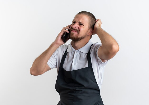 Unzufriedener junger hübscher Friseur, der Uniform trägt, die am Telefon spricht, seinen Kopf berührt und Seite betrachtet, die auf weißem Hintergrund lokalisiert wird