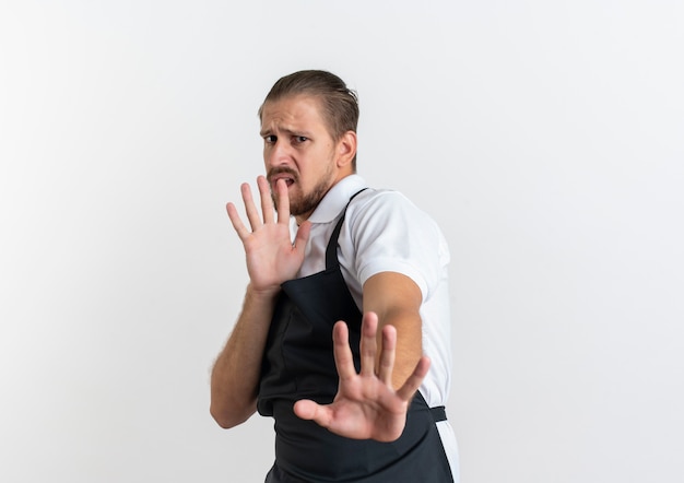 Unzufriedener junger hübscher Friseur, der Uniform trägt, streckt Hand in Richtung Kamera aus, die nicht isoliert auf weißem Hintergrund mit Kopienraum gestikuliert