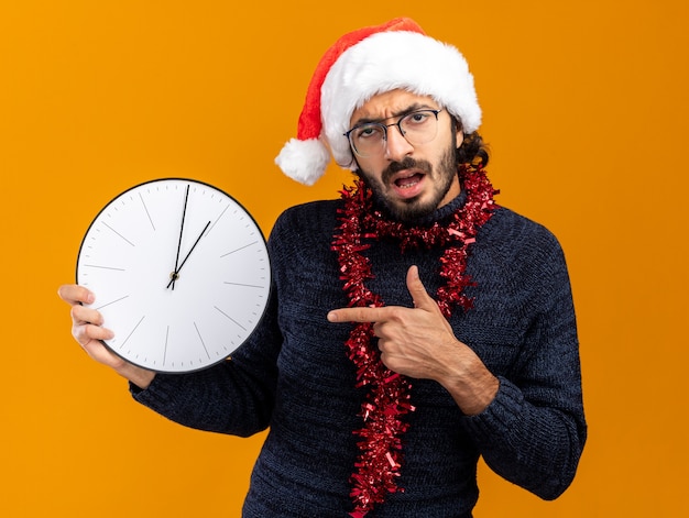 Unzufriedener junger hübscher Kerl, der Weihnachtsmütze mit Girlande am Hals hält und Punkte auf Wanduhr lokalisiert auf orange Hintergrund trägt