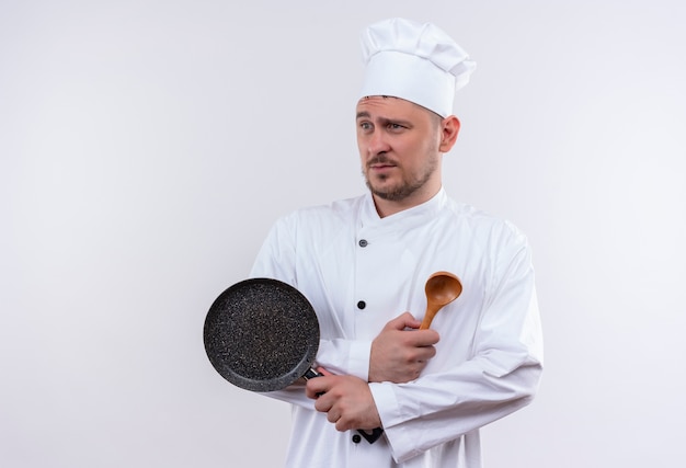 Unzufriedener junger hübscher Koch in der Kochuniform, die Bratpfanne und Löffel hält, die Seite lokalisiert auf weißem Raum betrachten