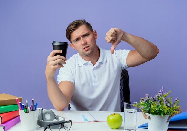 Unzufriedener junger hübscher männlicher Student, der am Schreibtisch mit Schulwerkzeugen sitzt, die Tasse Kaffee seinen Daumen nach unten lokalisiert auf Blau halten