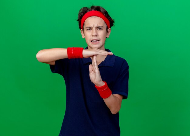 Unzufriedener junger hübscher sportlicher Junge, der Stirnband und Armbänder mit Zahnspangen trägt, die Kamera betrachten, die Zeitüberschreitungsgeste lokalisiert auf grünem Hintergrund mit Kopienraum tut
