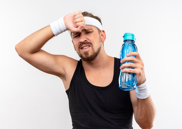 Unzufriedener junger hübscher sportlicher Mann, der Stirnband und Armbänder hält, die Wasserflasche mit Hand auf Kopf halten, lokalisiert auf weißem Raum