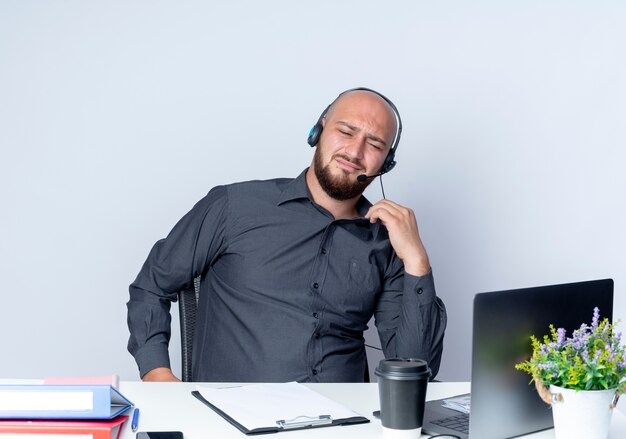Unzufriedener junger kahlköpfiger Callcenter-Mann, der Headset am Schreibtisch mit Arbeitswerkzeugen hält, die seinen Kragen halten, der lokalisiert auf weißem Hintergrund schaut