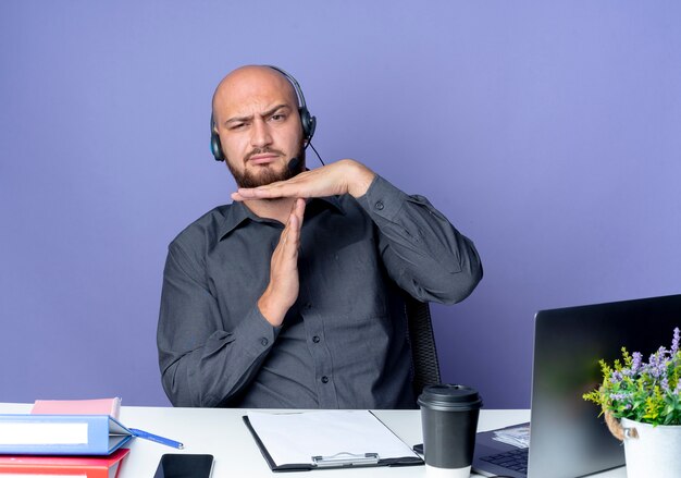 Unzufriedener junger kahlköpfiger Callcenter-Mann, der Headset sitzt am Schreibtisch mit Arbeitswerkzeugen, die Zeitüberschreitungsgeste tun, lokalisiert auf lila Hintergrund