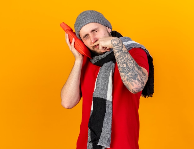 Unzufriedener junger kaukasischer kranker Mann, der Wintermütze und Schal trägt, wischt Nase mit Fingern ab und setzt Kopf auf Wärmflasche lokalisiert auf orange Wand mit Kopienraum