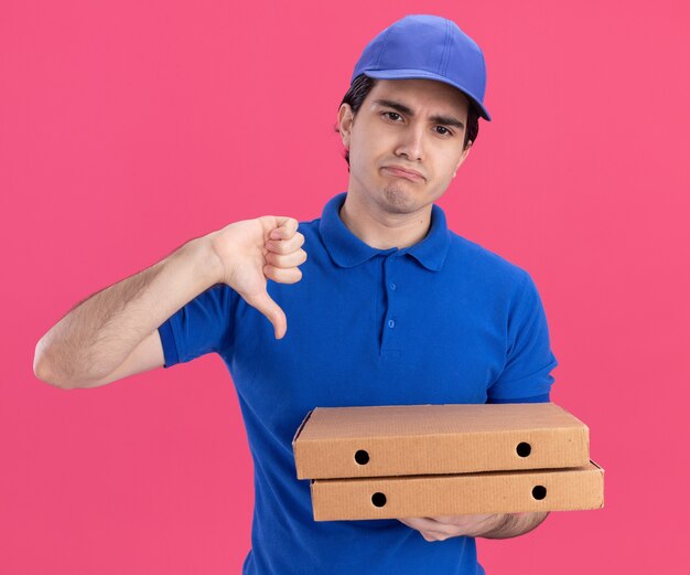 Unzufriedener junger kaukasischer Lieferbote in blauer Uniform und Mütze, die Pizzapakete hält, die gerade mit dem Daumen nach unten schauen