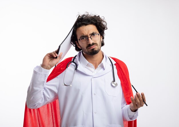 Unzufriedener junger kaukasischer Superheldenmann in der optischen Brille, die Arztuniform mit rotem Umhang und mit Stethoskop um Hals trägt, setzt Klemmbrett auf Kopf und hält Bleistift auf weißer Wand