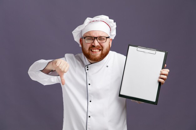 Unzufriedener junger Koch mit einheitlicher Brille und Mütze, die Klemmbrett zeigt und in die Kamera schaut, die den Daumen nach unten zeigt, isoliert auf violettem Hintergrund