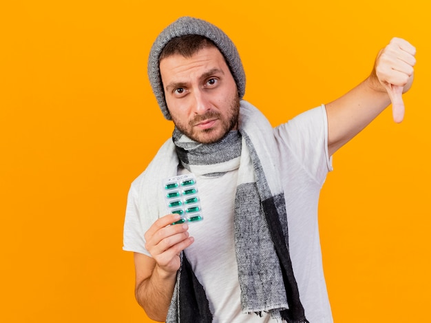 Unzufriedener junger kranker Mann, der Wintermütze und Schal hält, die Pillen halten und Daumen unten lokalisiert auf gelbem Hintergrund zeigen