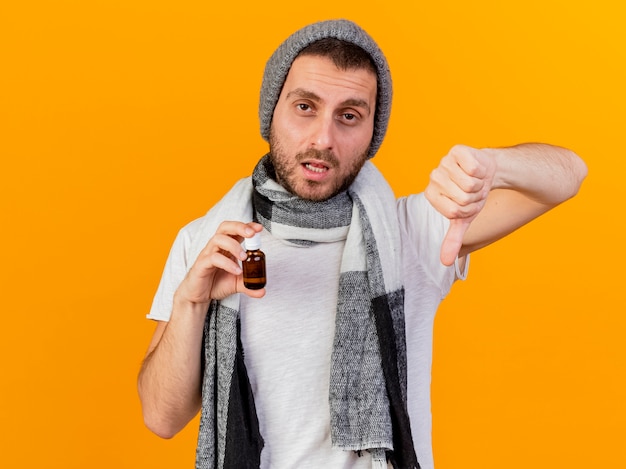 Unzufriedener junger kranker Mann, der Wintermütze und Schal hält Medizin in der Glasflasche zeigt Daumen unten auf gelbem Hintergrund