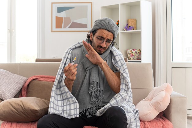 unzufriedener junger kranker Mann in optischer Brille, eingewickelt in Plaid mit Schal um den Hals, der eine Wintermütze trägt und die Spritze auf der Couch im Wohnzimmer sieht