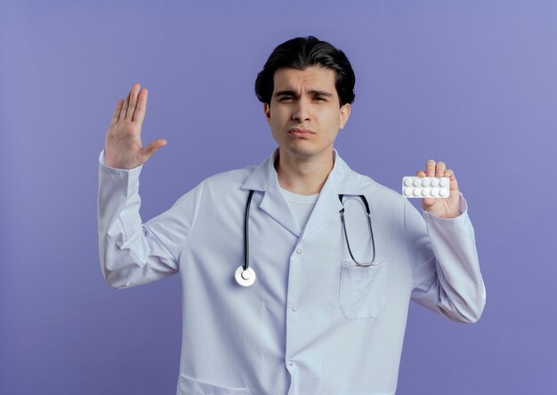 Unzufriedener junger männlicher Arzt, der medizinisches Gewand und Stethoskop trägt, die Packung der Tabletten tun, die Stoppgeste zeigen, die auf lila Wand lokalisiert werden