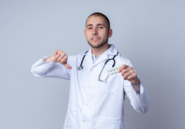 Unzufriedener junger männlicher Arzt, der medizinisches Gewand und Stethoskop um seinen Hals trägt, Packung der medizinischen Tabletten hält und Daumen unten lokalisiert auf weißem Hintergrund zeigt