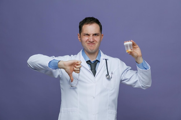 Unzufriedener junger männlicher Arzt mit medizinischem Gewand und Stethoskop um den Hals, der in die Kamera blickt und Messbehälter mit Pillen darin und Daumen nach unten isoliert auf violettem Hintergrund zeigt