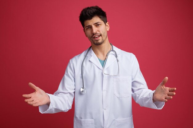 Unzufriedener junger männlicher Arzt mit medizinischer Uniform und Stethoskop um den Hals, der in die Kamera blickt und leere Hände isoliert auf rotem Hintergrund zeigt