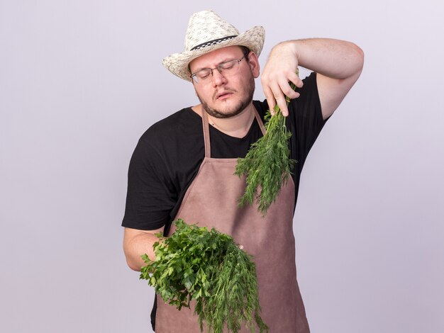 Unzufriedener junger männlicher Gärtner, der Gartenhut hält und Dill mit Koriander lokalisiert auf weißer Wand hält