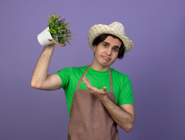 Kostenloses Foto unzufriedener junger männlicher gärtner in der uniform, die gartenhut erhöht und auf blume im blumentopf zeigt