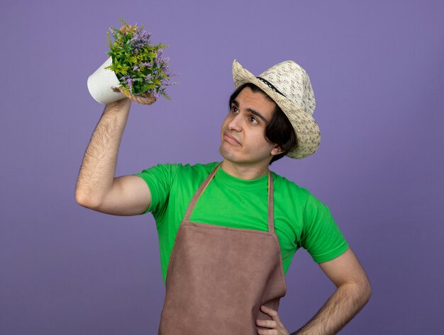 Unzufriedener junger männlicher Gärtner in der Uniform, die Gartenhut trägt, der Blume im Blumentopf anhebt und Hand auf Hüfte setzt, die auf Purpur lokalisiert