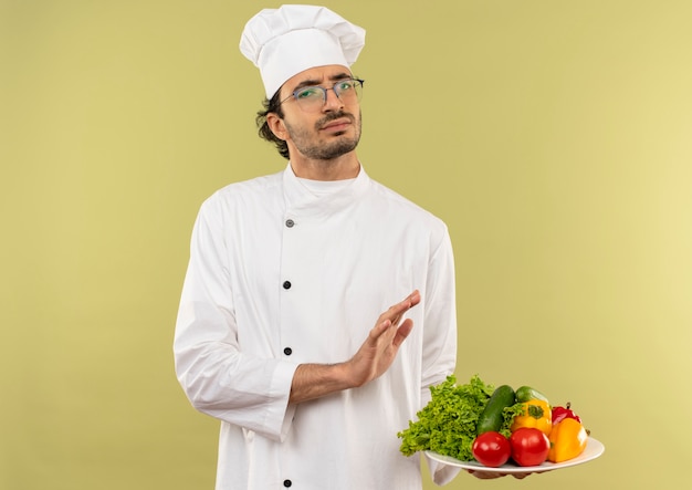 unzufriedener junger männlicher Koch, der Kochuniform und Gläser trägt, die Gemüse auf Teller halten und Stoppgeste zeigen